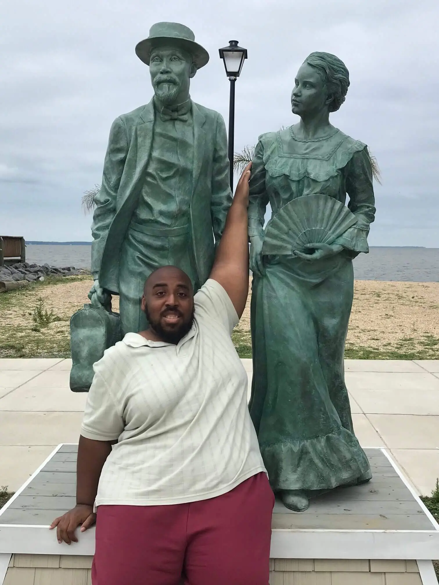 Man posing with statues of a man and woman at a beach.