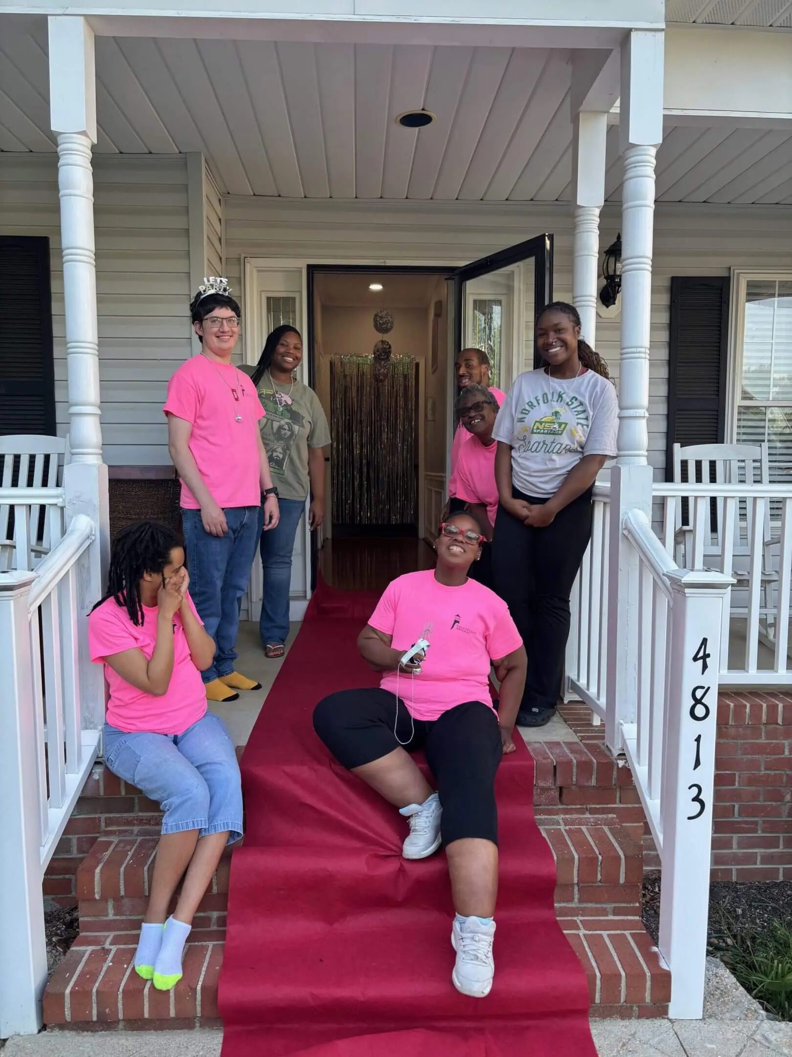 Five people in pink shirts posing on a porch with a white house background.